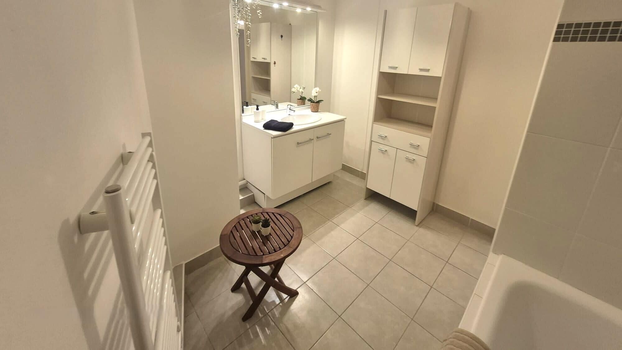 A bathroom with a white sink, mirror, and cabinets, a small wooden stool, and a tiled floor.