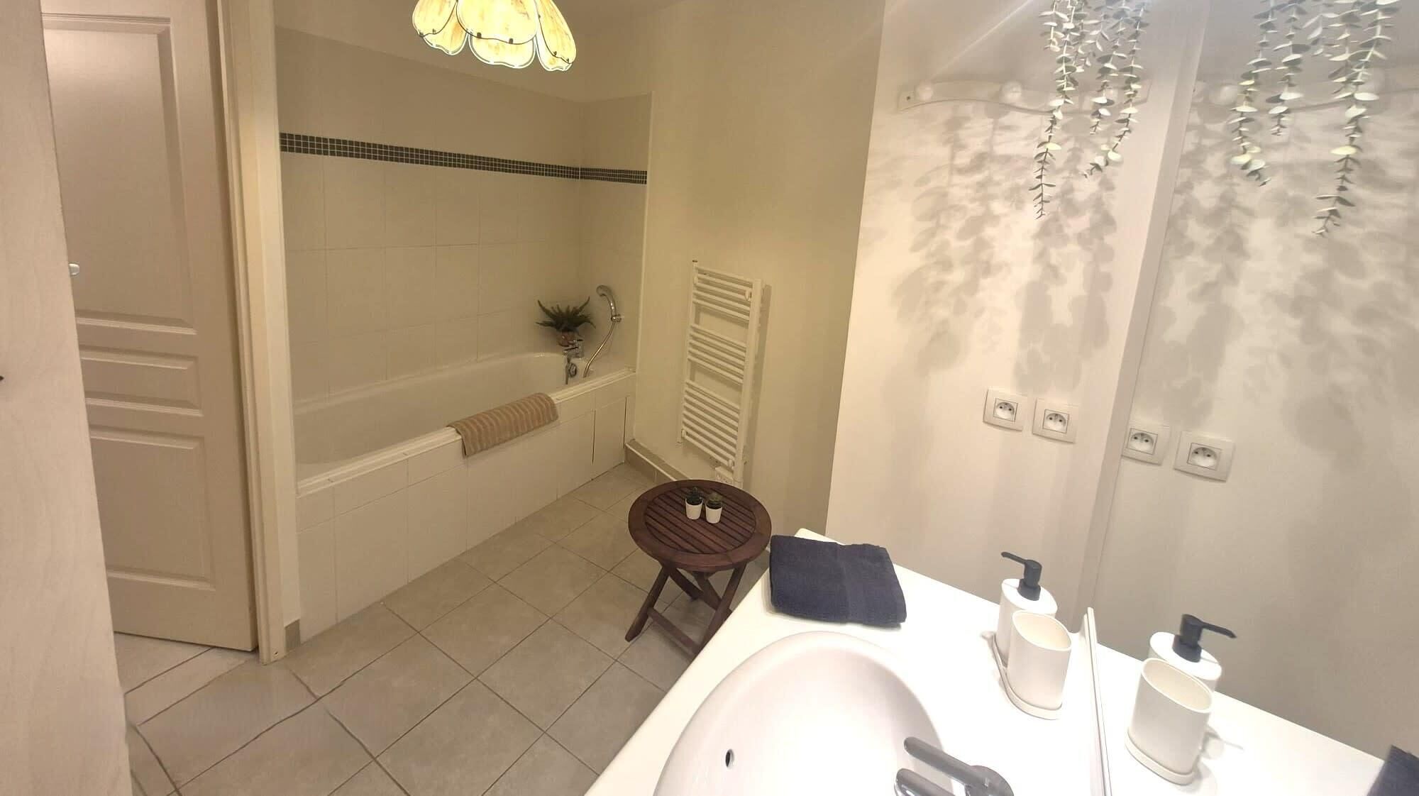 A bathroom with a bathtub, a round wooden stool, a sink with toiletries, and a towel rack.
