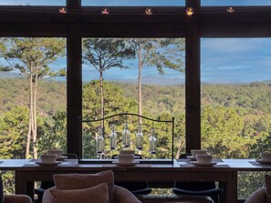 Ein Essbereich mit Blick auf eine bewaldete Landschaft durch große Fenster, einer Metallleuchte und einem Tisch, der mit Schüsseln und Tellern gedeckt ist.