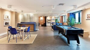 A modern recreational room with a pool table, blue chairs, a dining area, and a kitchen with a blue backsplash. - Student Only Hollis Wharf (Bath)