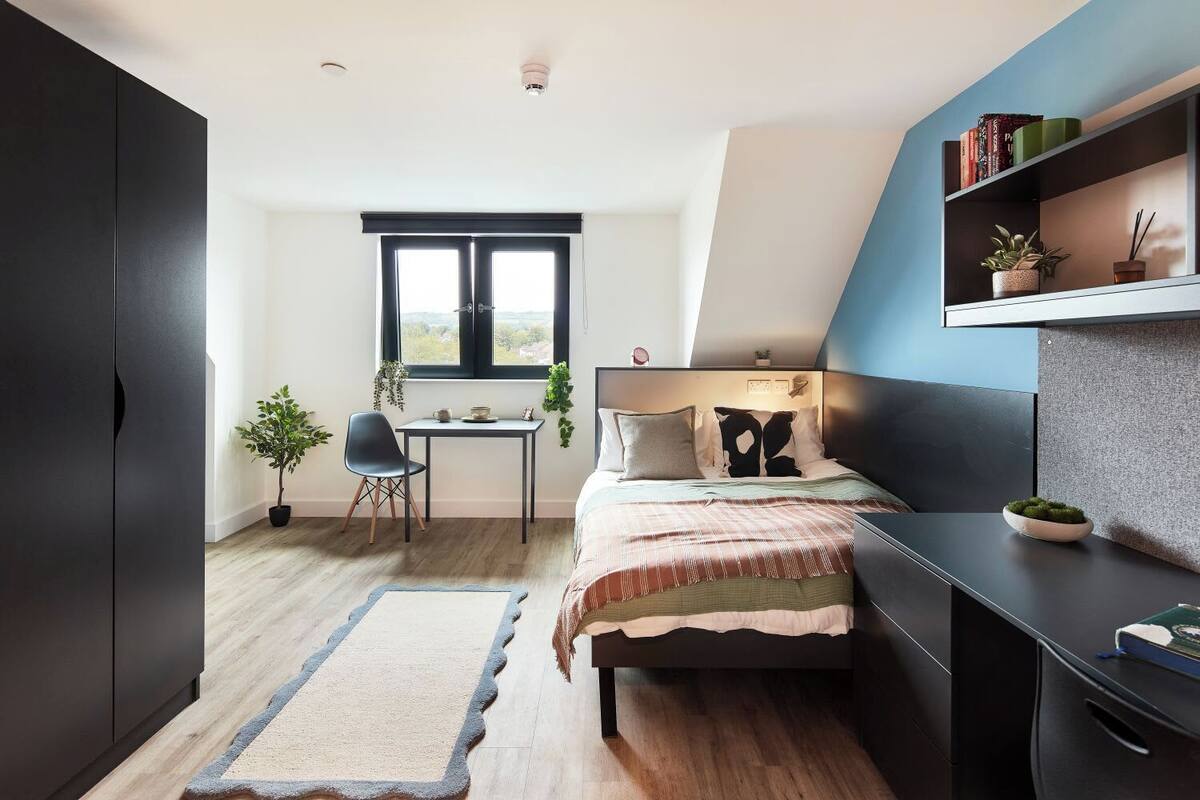 A bedroom with a bed, a desk, and a chair. There is a window with a view of the outside.