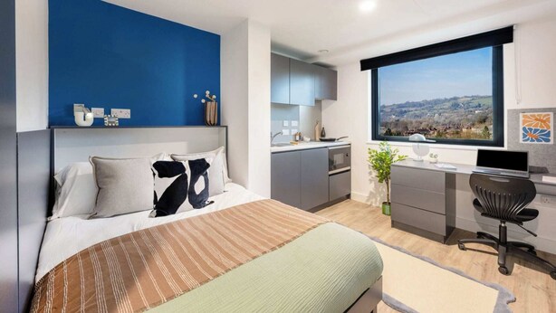 A modern bedroom with a large bed, a desk, and a view of a landscape through a window. - Student Only Hollis Wharf (Bath)