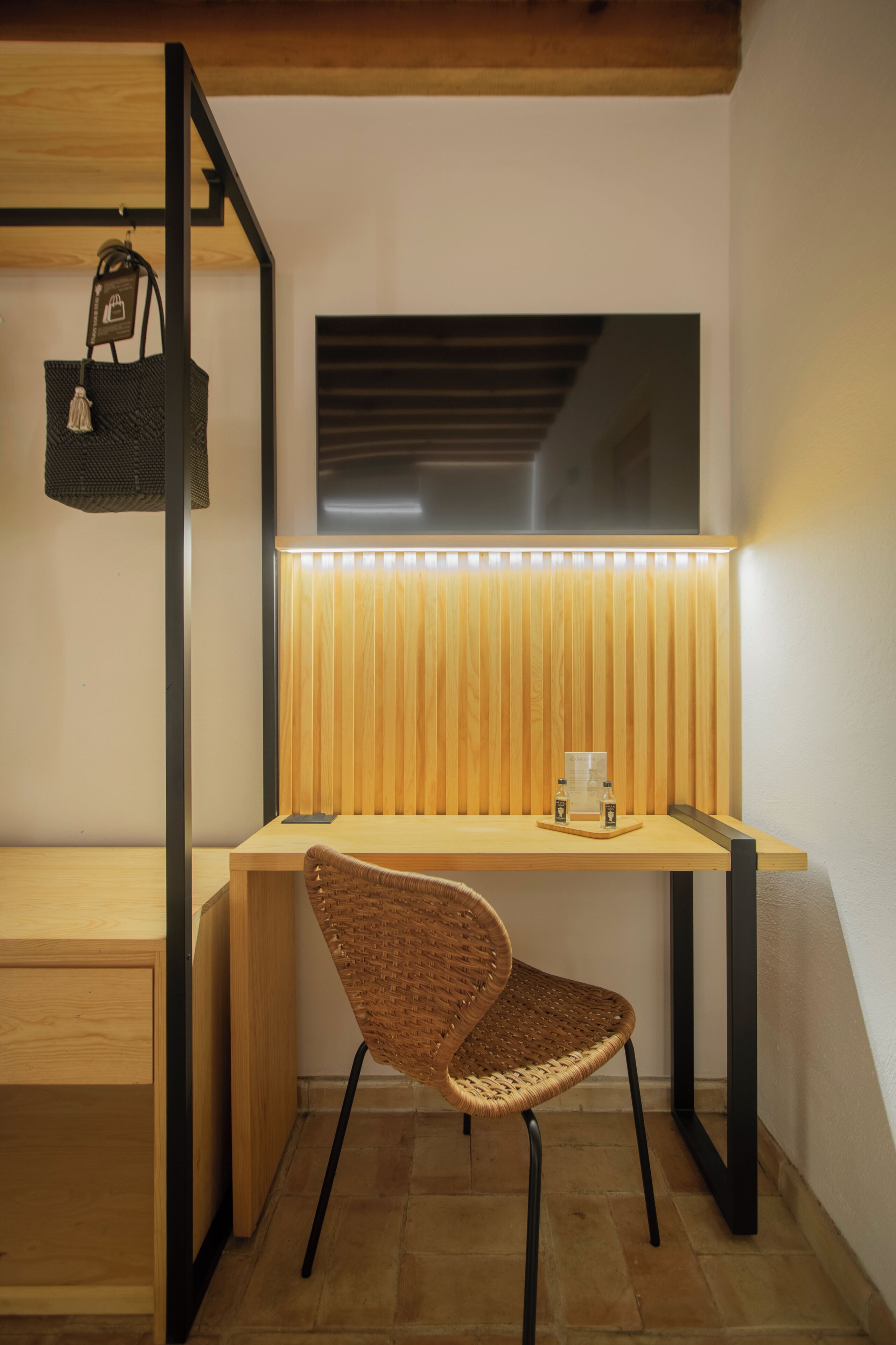 Un espacio de trabajo moderno con un escritorio de madera, una silla tejida, un bolso colgado y una lámpara de pared.