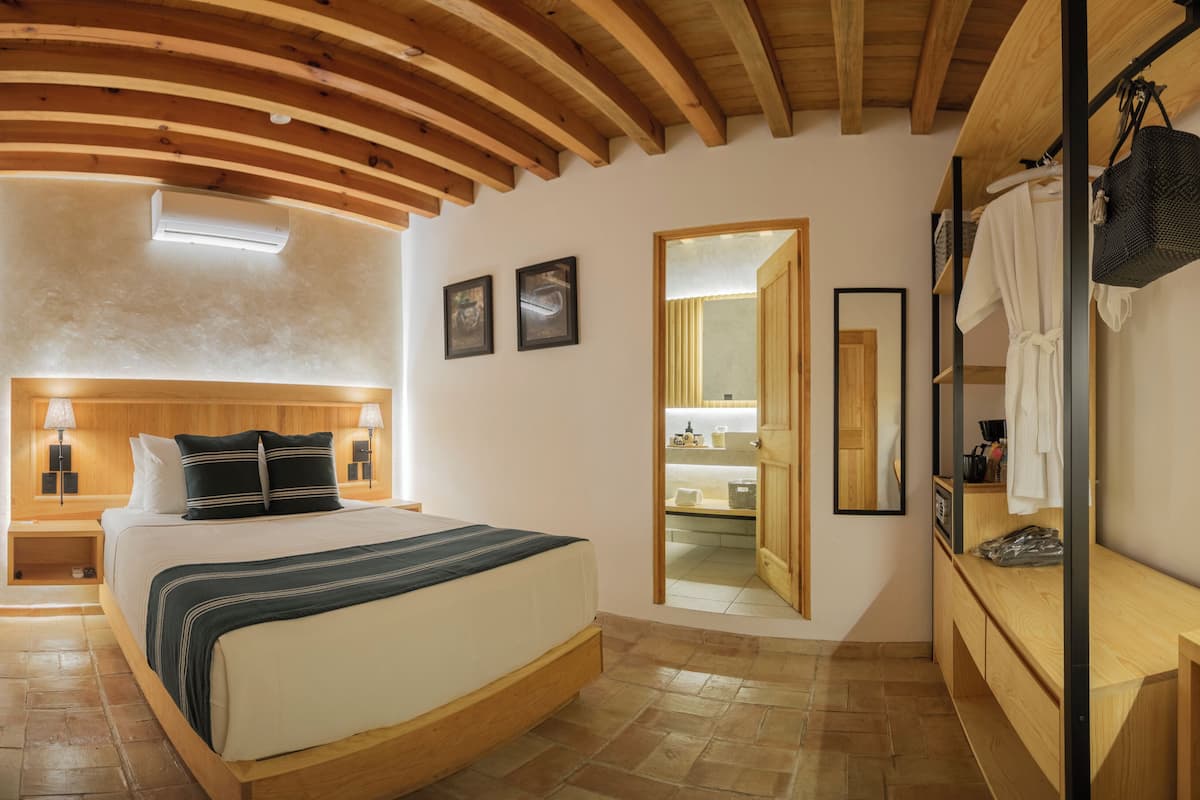 Un dormitorio con techo de madera, una cama con cabecera, una mesita de noche, una lámpara, un espejo y un banco de madera.