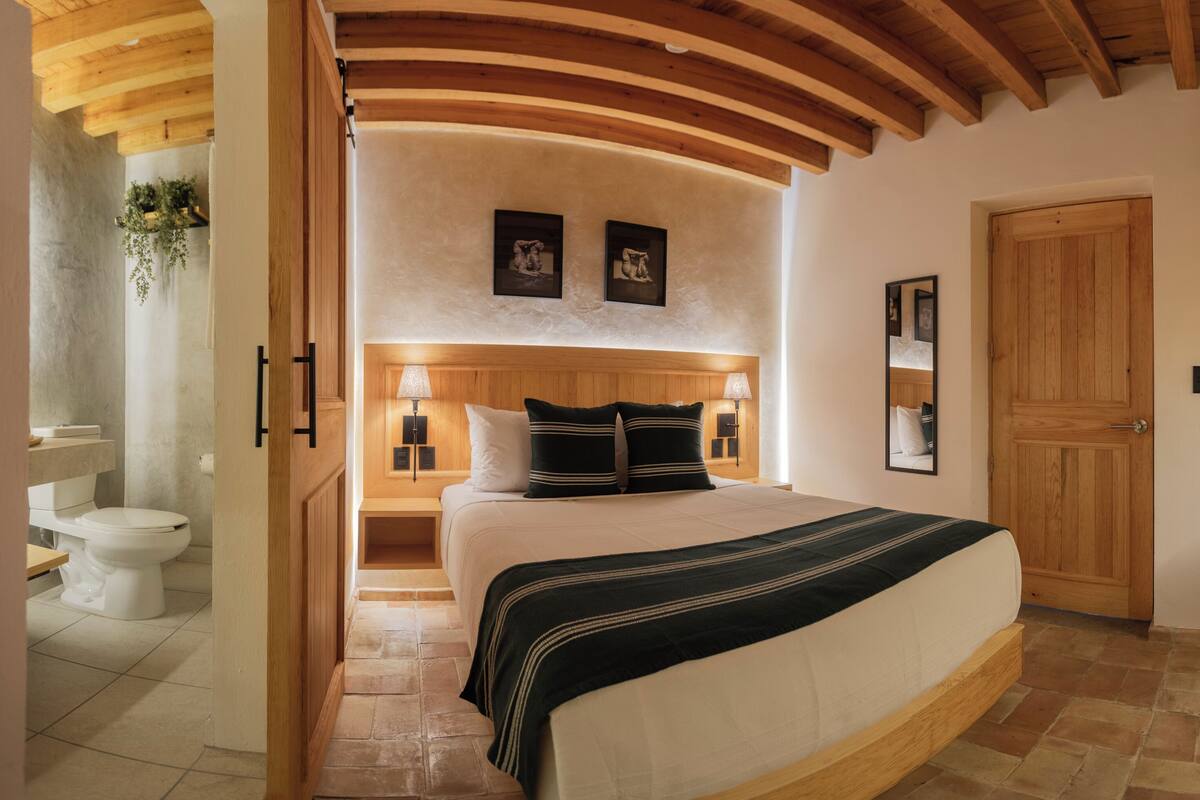 Un dormitorio con cama, vigas de techo de madera, puerta de madera, una silla y una mesita.
