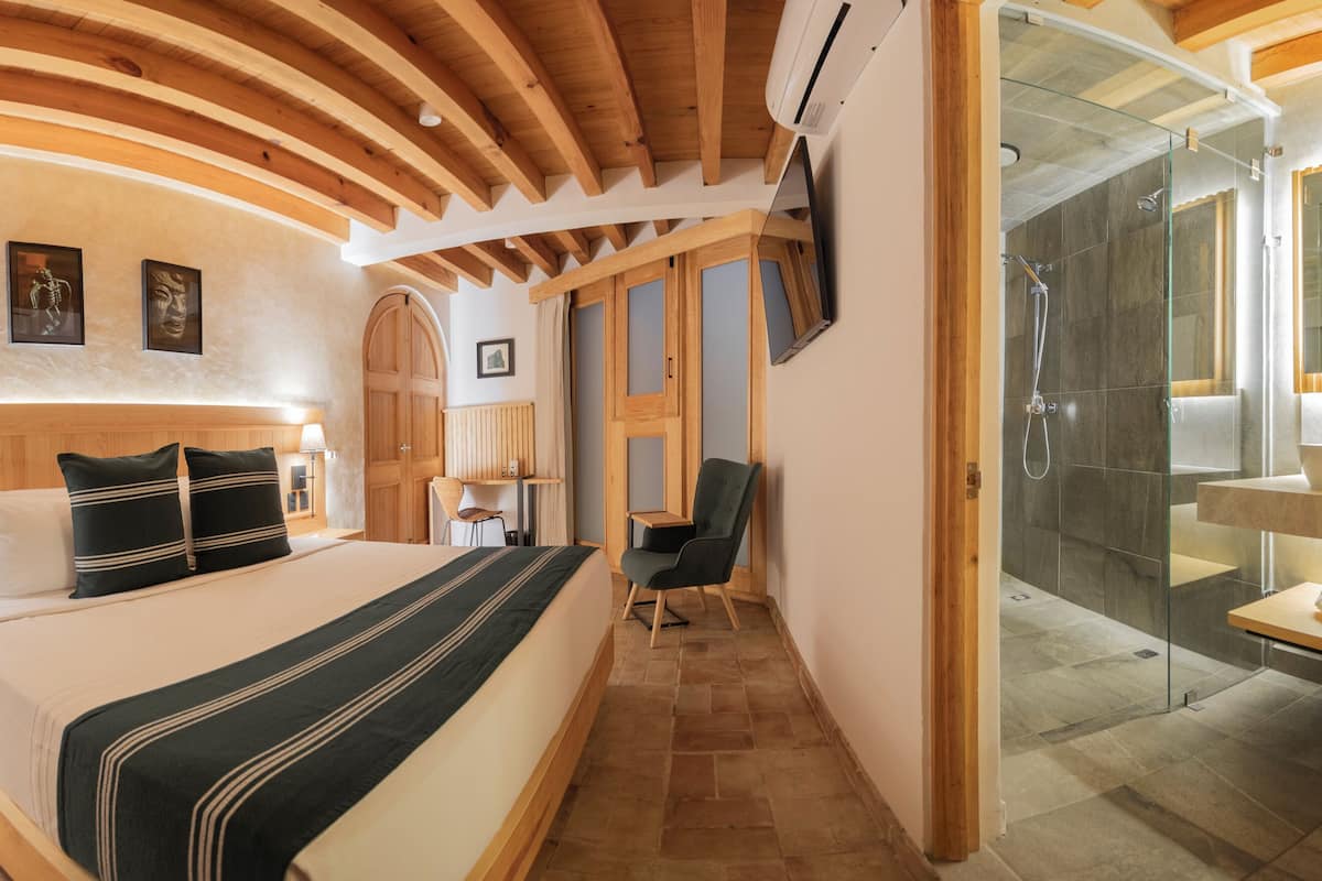 Una habitación de hotel moderna con una cama grande, vigas de madera en el techo, una ducha de vidrio y una pequeña mesa con una silla.