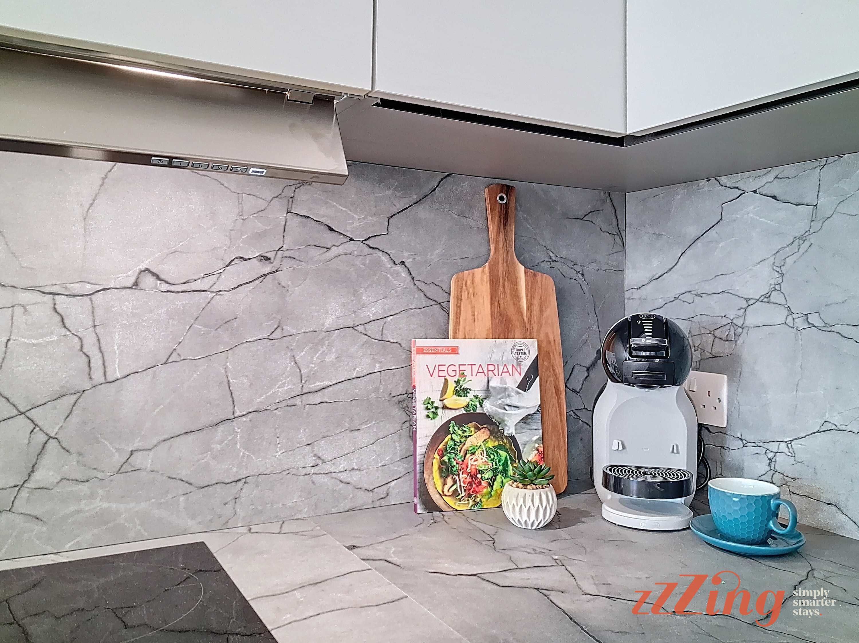 A kitchen with a marble backsplash, a wooden cutting board, a kettle, a cup, and a book titled "VEGETARIAN ESSENTIALS".