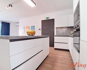 A modern kitchen with white cabinets, a black countertop, and a marble backsplash. There is a bowl of fruit on the counter and a door with an exit sign above it. - Balcony Apartment in Charming Malta Village (Ħal Għaxaq)