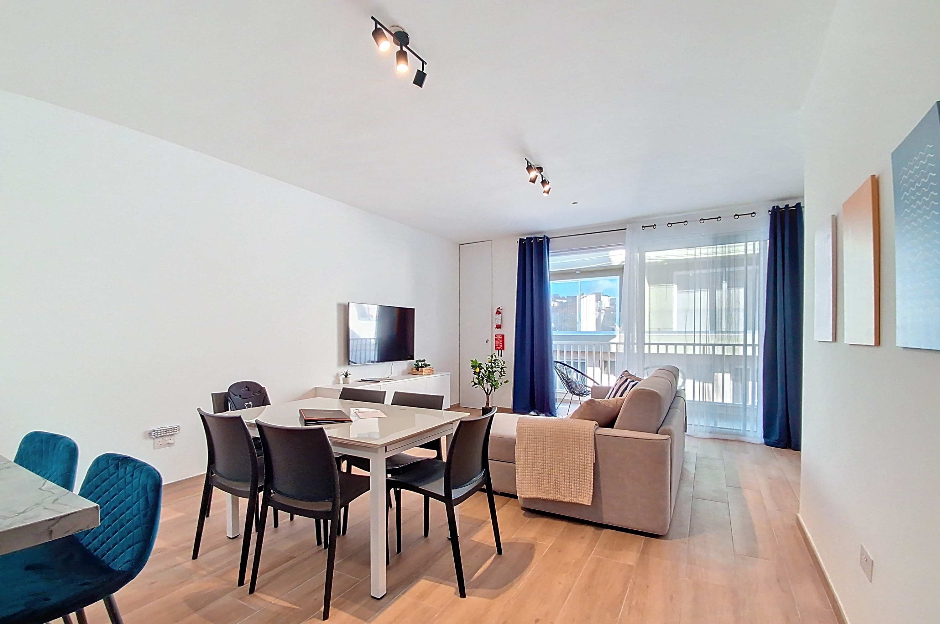 Una sala moderna con comedor, un sofá, un televisor y un balcón con cortinas.