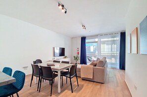 A modern living room with a dining area, a sofa, a TV, and a balcony with curtains. - Balcony Apartment in Charming Malta Village (Ħal Għaxaq)