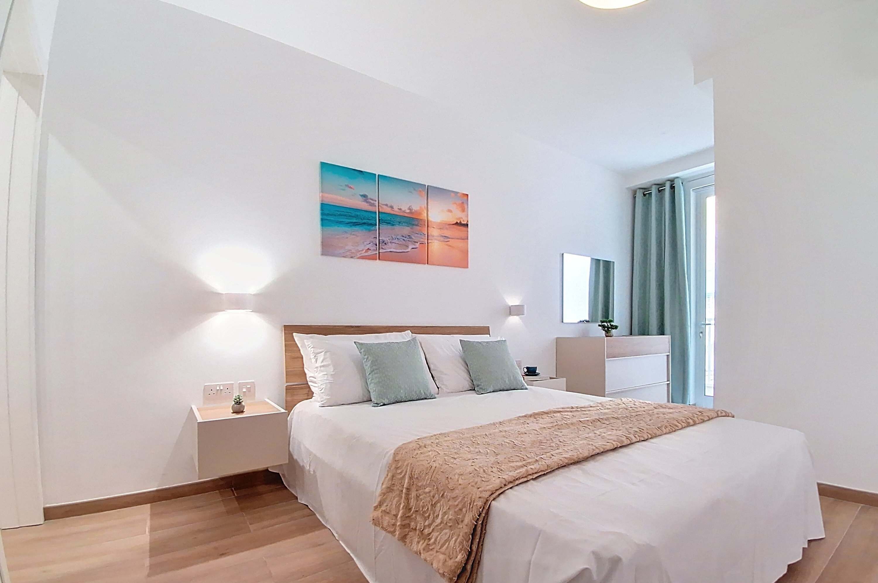Et moderne hotelværelse med en stor seng, et natbord, en lampe fastgjort til væggen og et maleri af en solnedgang over havet.