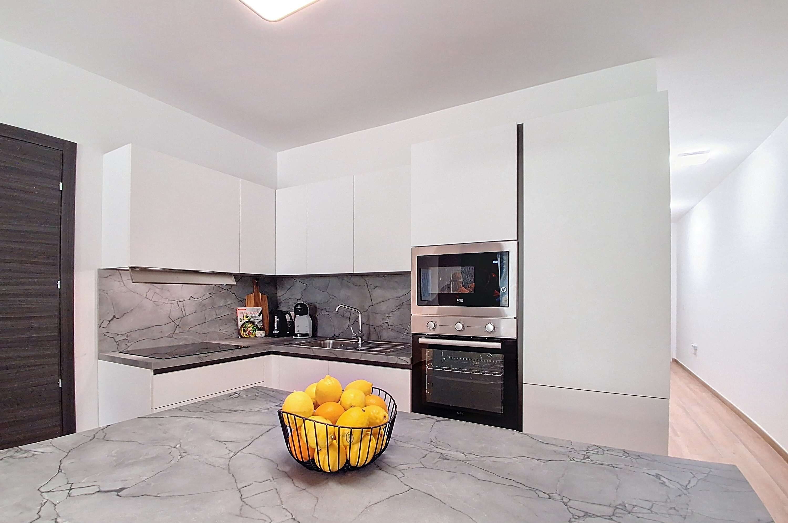 A modern kitchen with white cabinets, a marble countertop, a built-in oven, and a bowl of lemons on the island.