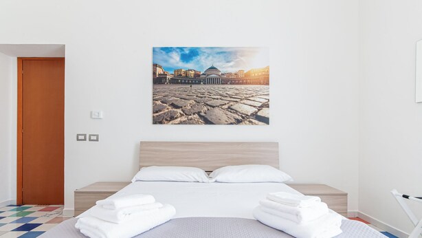 A hotel room with a large bed, neatly folded towels, a wooden headboard, and a cityscape painting on the wall. - Apartment in Plebiscito int 11 (Napoli)