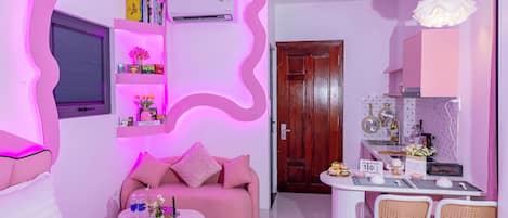 A modern room with a pink sofa, a small round table, and a kitchenette area with a wooden door.