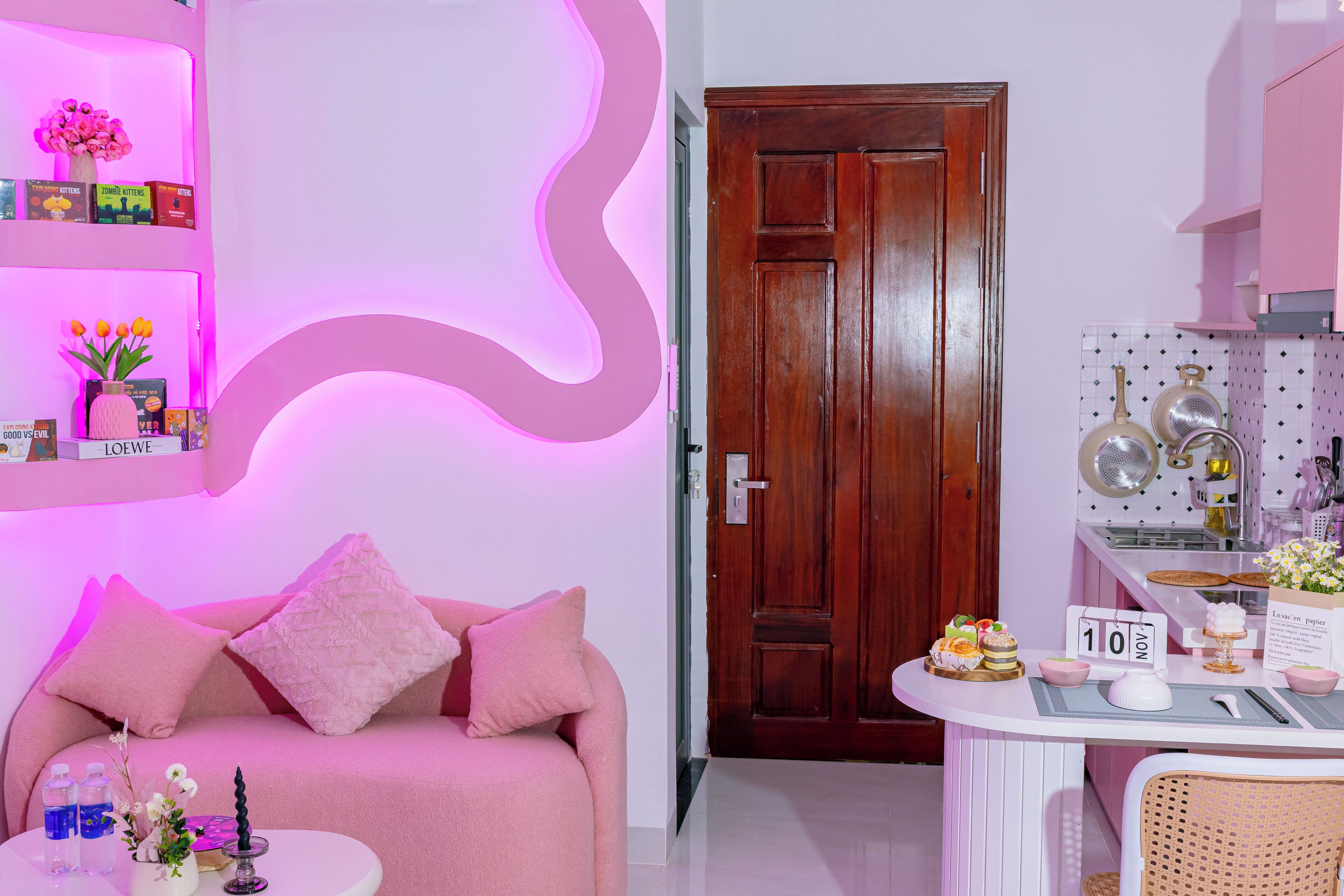 A room with a pink sofa, a small table with decorative items, a wooden door, and a kitchen area with pots and a calendar.