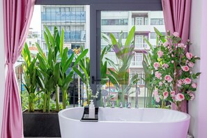 A modern bathroom with a large bathtub, potted plants, and a view of a cityscape through a window. - ORI HOME (Ho Chi Minh City)
