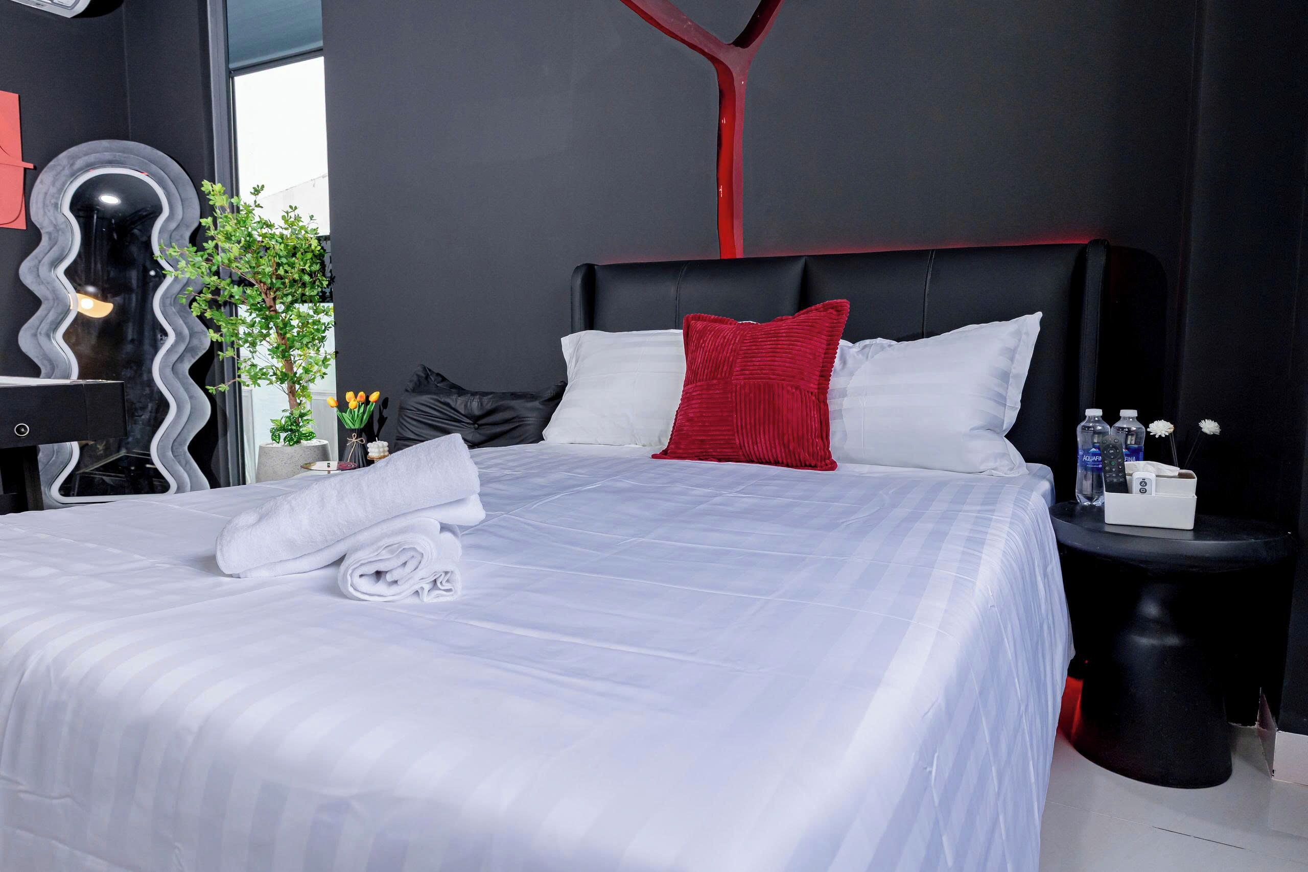 A modern hotel room with a large bed, a red accent pillow, a black nightstand, and a mirror with a wavy frame.