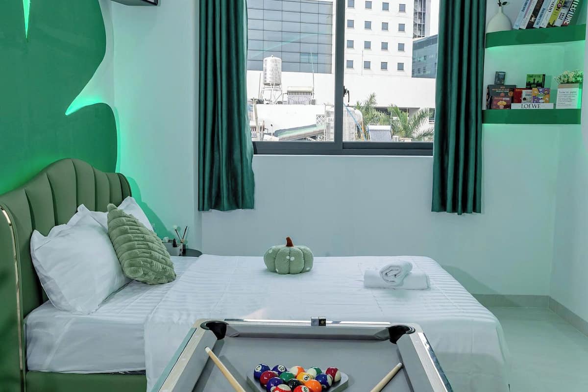 A room with a pool table, a bed with green and white bedding, a window with curtains, and a view of buildings outside.