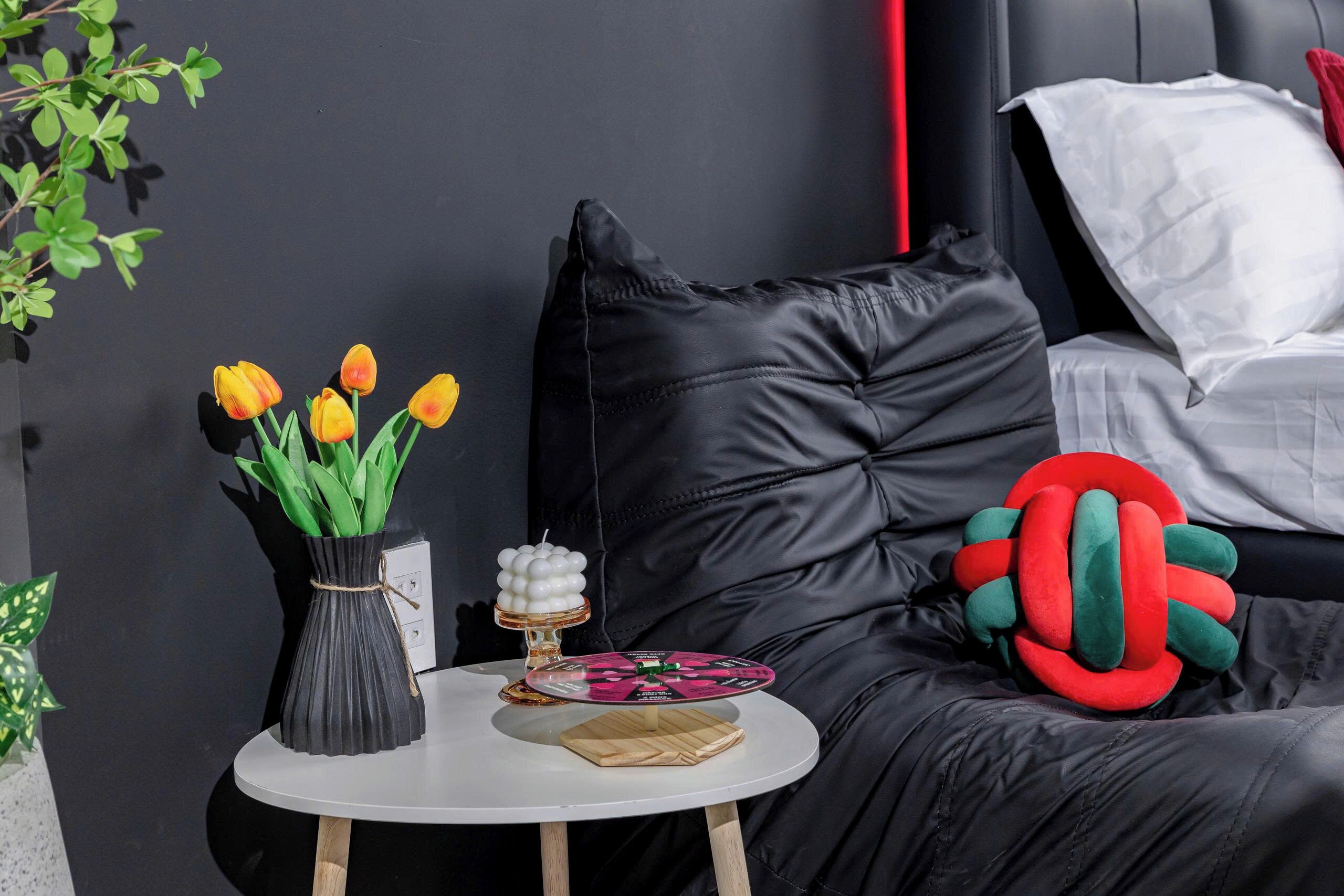 A modern bedroom with a black leather headboard, a white pillow, a red and green plush toy, a black vase with yellow tulips, and a small round table with a white surface.