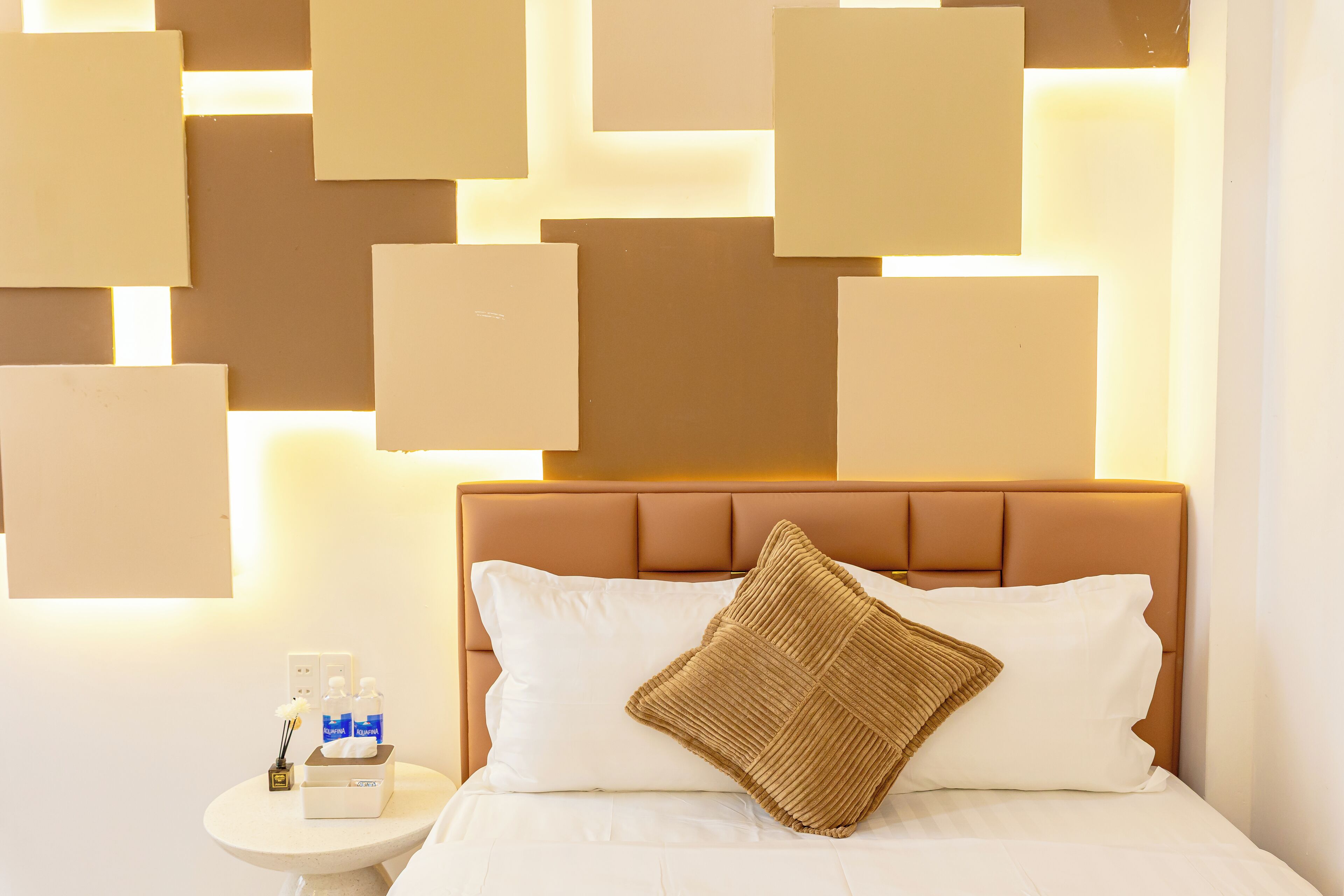 A neatly made bed with a white pillow and a textured pillow, a bedside table with a bottle of lotion and a box of tissues, and a wall with geometric light panels.