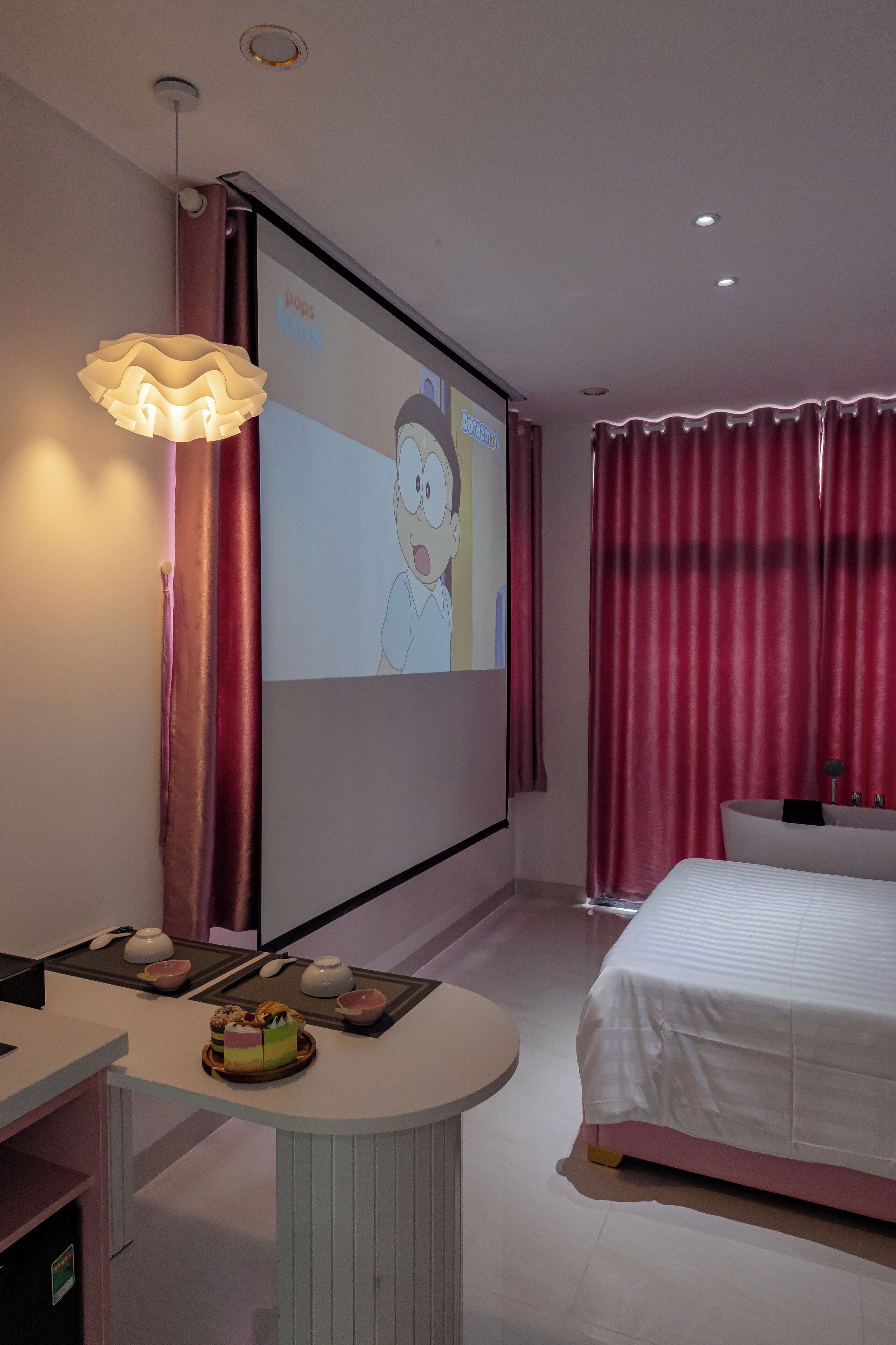 A hotel room with a bed, a desk with a laptop, a projector screen, and a red curtain.