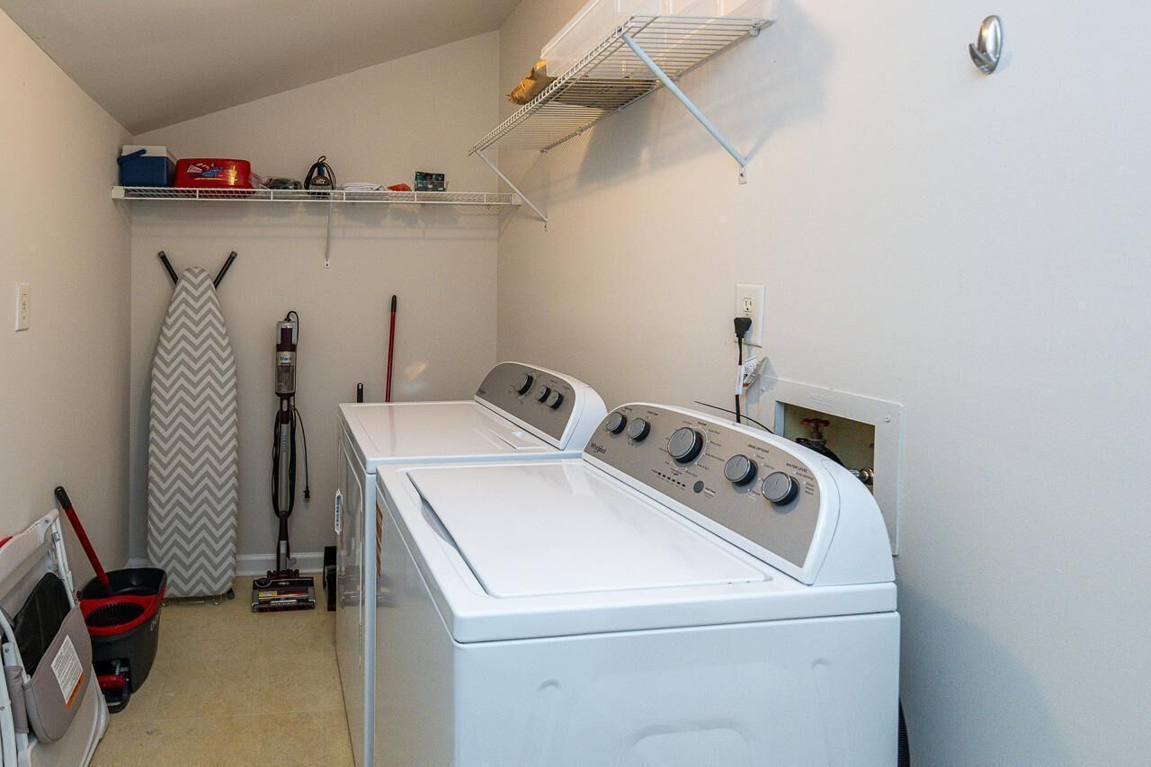 Une buanderie équipée de deux machines à laver et deux sèche-linge superposés, d’une chaise pliante, d’un balai et d’une étagère contenant divers objets.