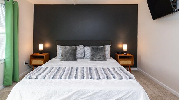 Une chambre à coucher avec un grand lit, deux tables de chevet avec des lampes, une tête de lit sombre et un téléviseur fixé au mur.