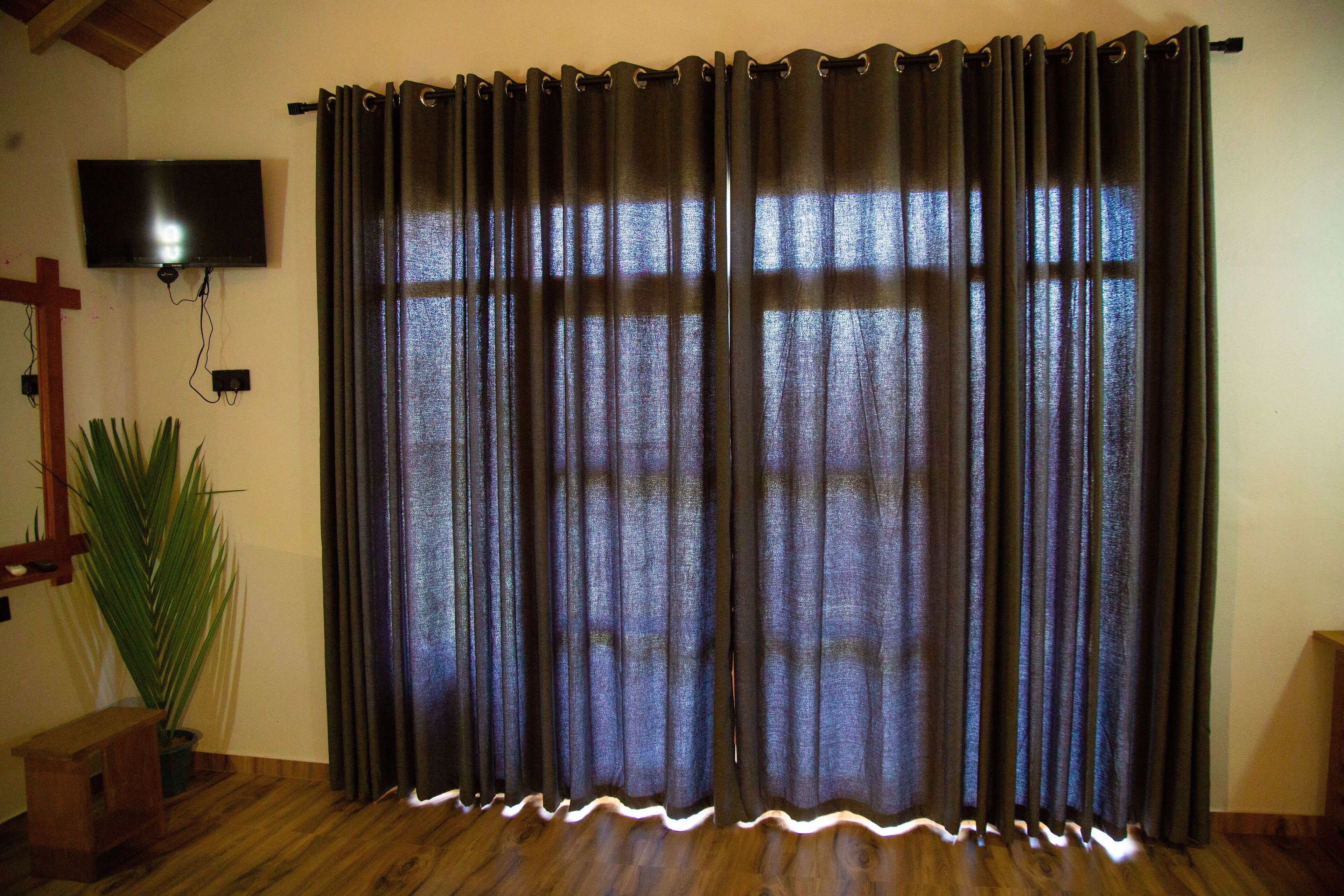 A room with a large window covered by dark curtains, a flat-screen TV mounted on the wall, and a wooden floor.