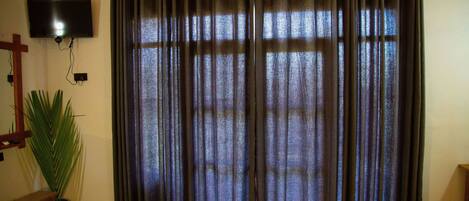 A room with a large window covered by dark curtains, a flat-screen TV mounted on the wall, and a wooden floor.
