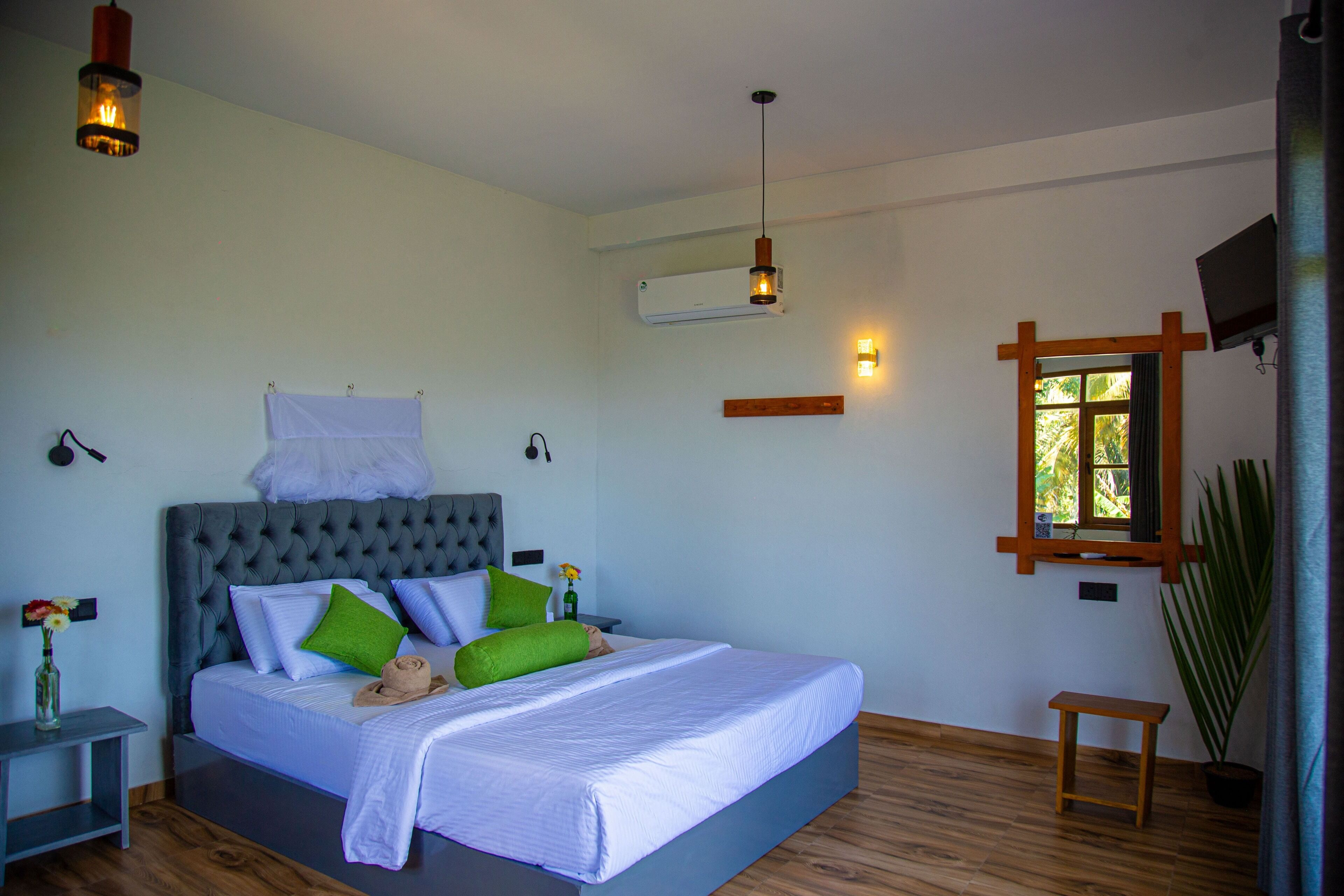 A bedroom with a large bed, a bedside table, a nightstand, a potted plant, and a window with a view of greenery.