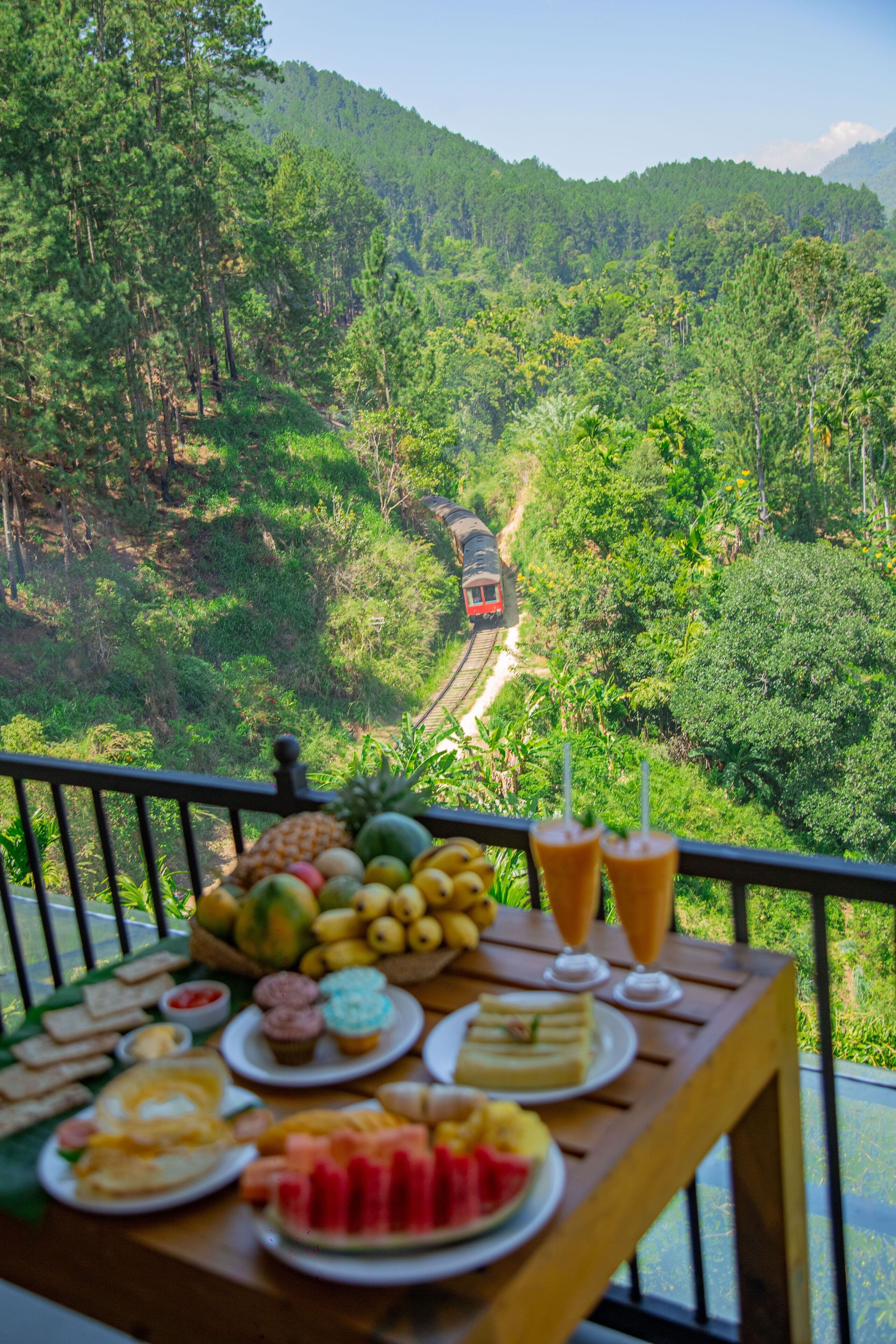 A table with various fruits and pastries, a train in the background, and a lush green forest.