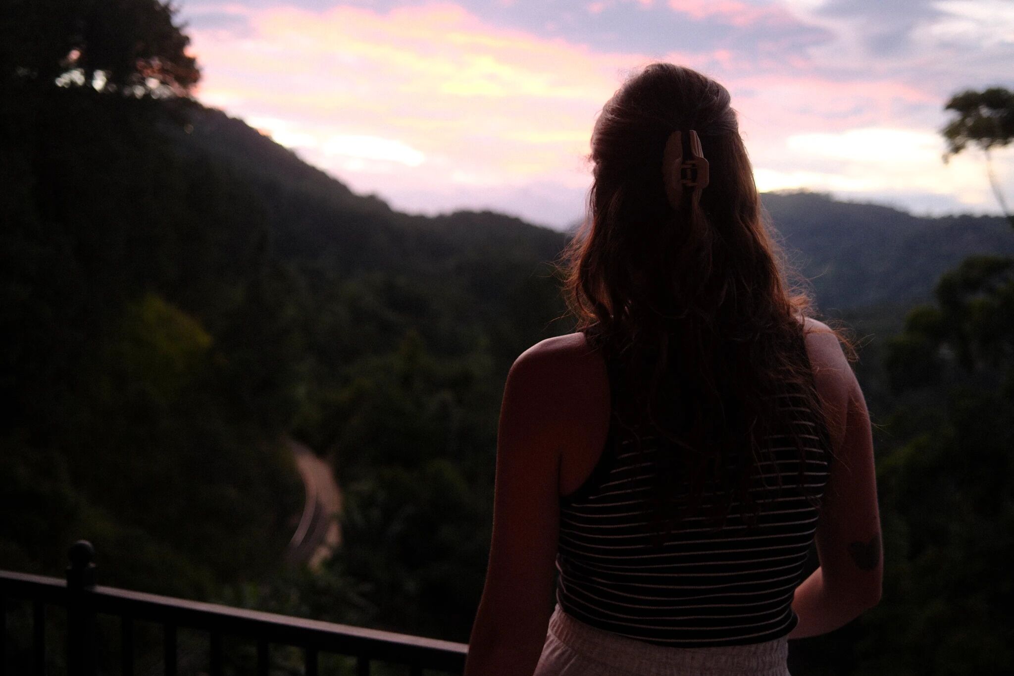Eine Person steht auf einem Balkon mit Blick auf eine gebirgige Landschaft und eine kurvenreiche Straße.