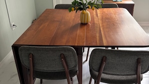 Un coin repas avec une table en bois, deux chaises rembourrées, un vase de citrons et une fenêtre avec des stores.