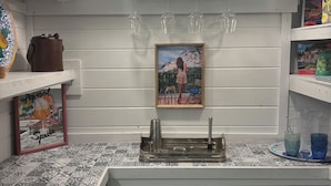 Une cuisine avec un mur recouvert de carreaux, un évier, des verres à vin suspendus, un tableau encadré et une assiette décorative.