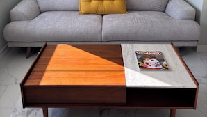 Un salon avec un canapé gris, un coussin de couleur moutarde, une table basse en bois et un tableau encadré représentant un paysage côtier.
