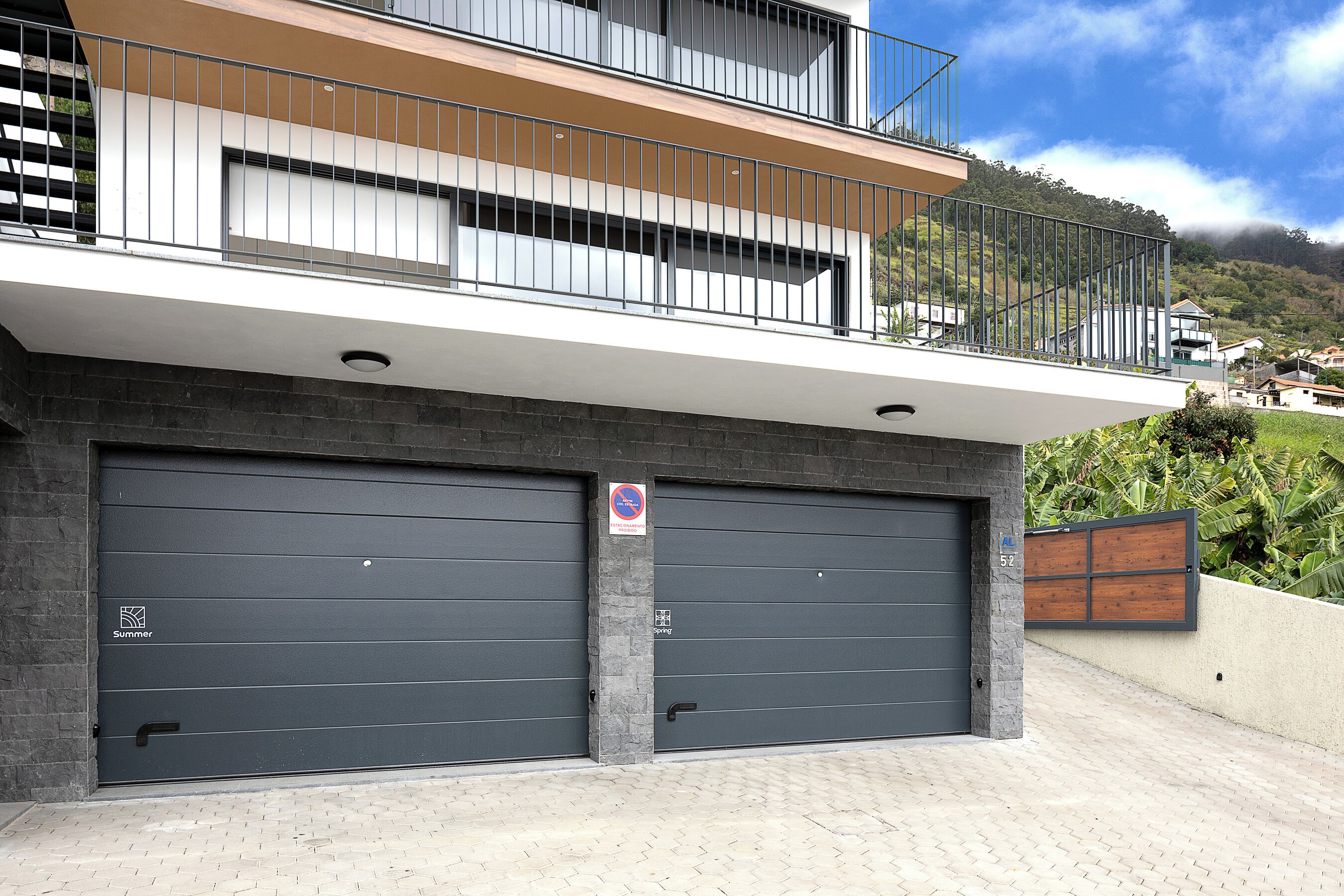 A modern building with two garage doors, a balcony, and a view of a hilly landscape.