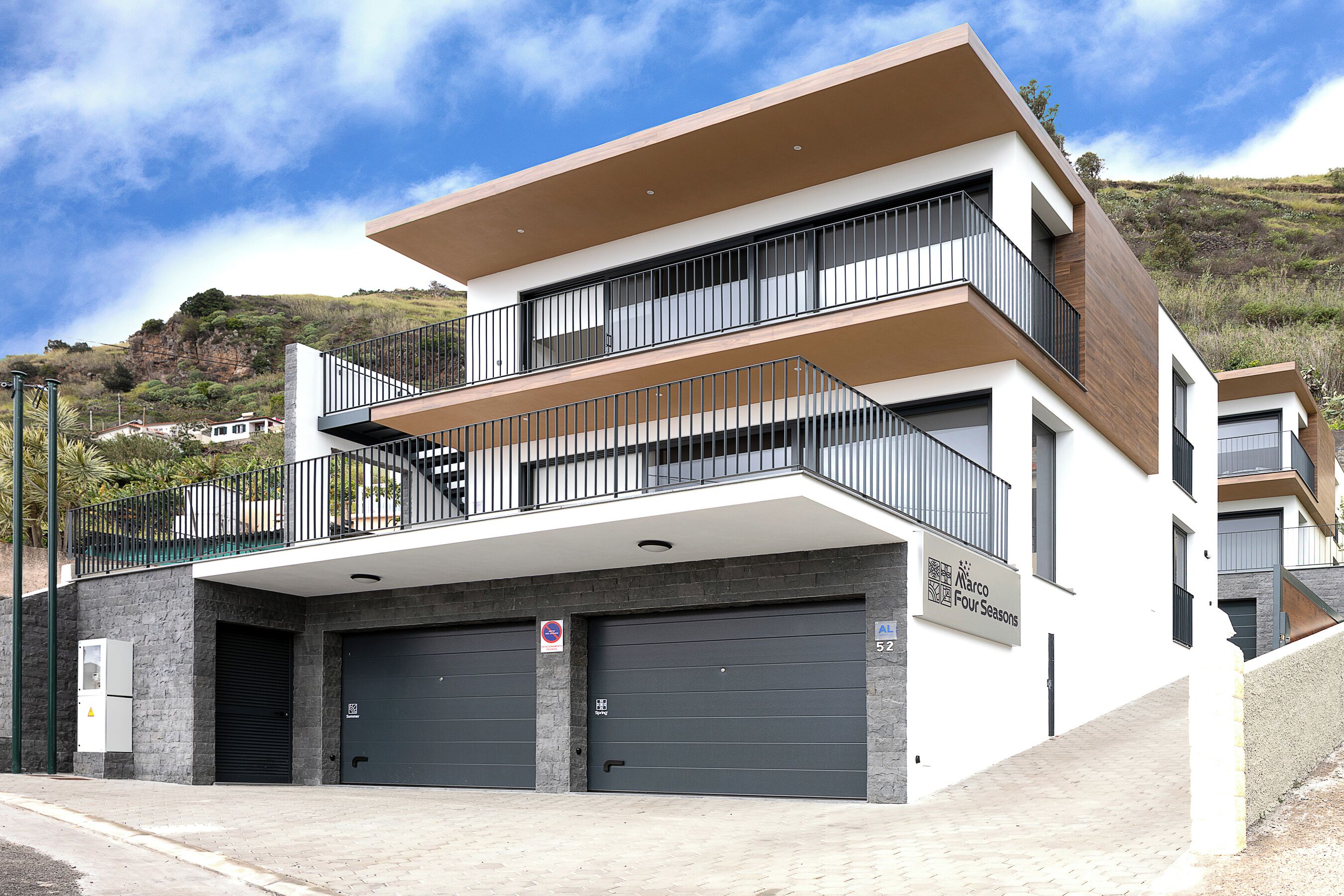 Ein modernes, mehrstöckiges Gebäude mit Flachdach, zwei Garagentoren und einem Balkon mit Metallgeländern.