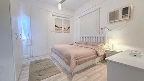 A bedroom with a bed, bedside table, and a nightstand. There is a window with curtains, a wall-mounted air conditioner, and a radiator. - Maskn Al Bader (Madinah)