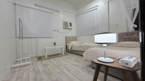A bedroom with two beds, a bedside table with a lamp, a small bedside table with a plant, and a window with curtains. - Maskn Al Bader (Madinah)