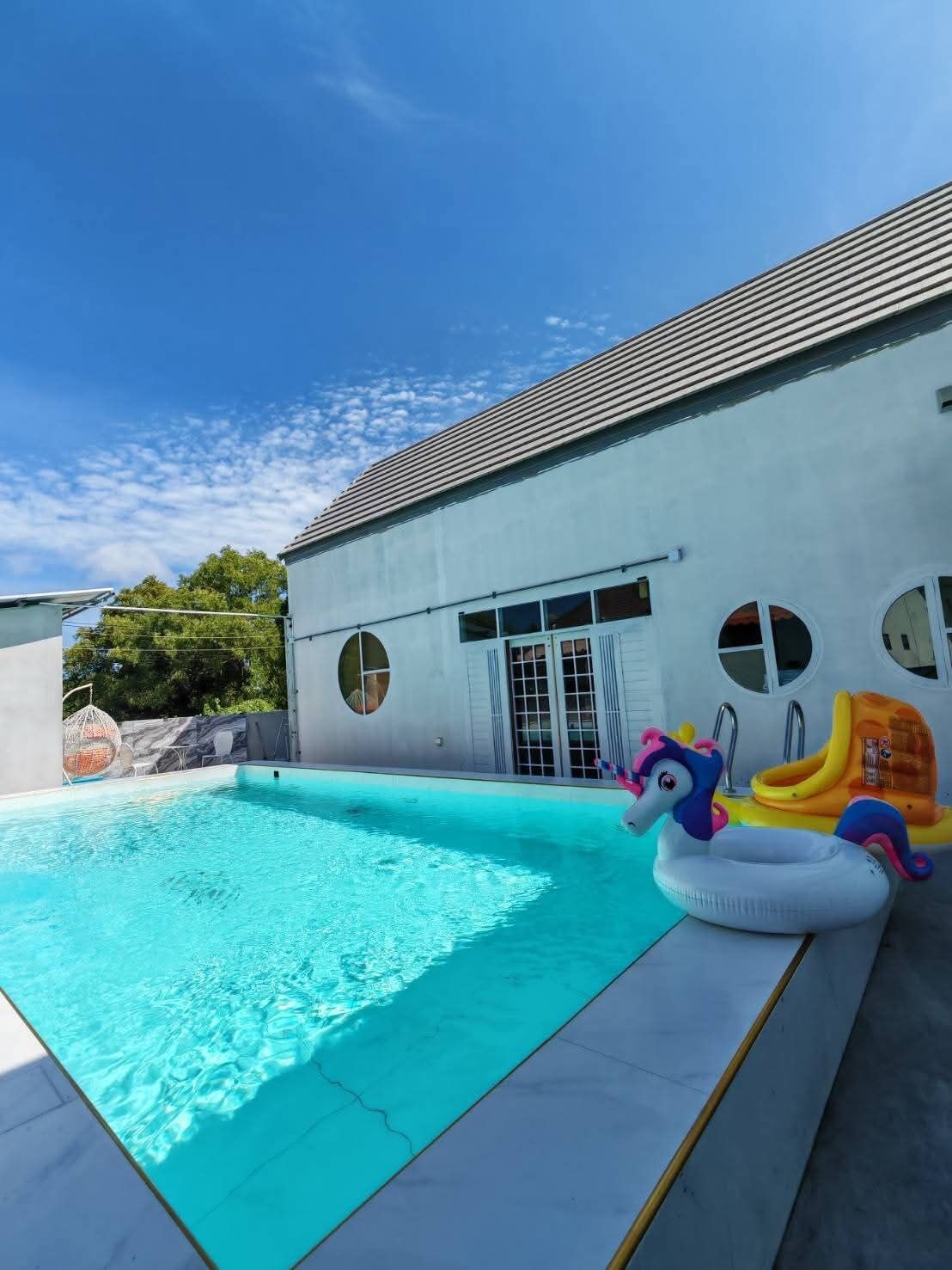 Ein Schwimmbecken mit aufblasbaren Spielzeugen, ein weißes Gebäude mit Rundfenstern und ein strahlend blauer Himmel.