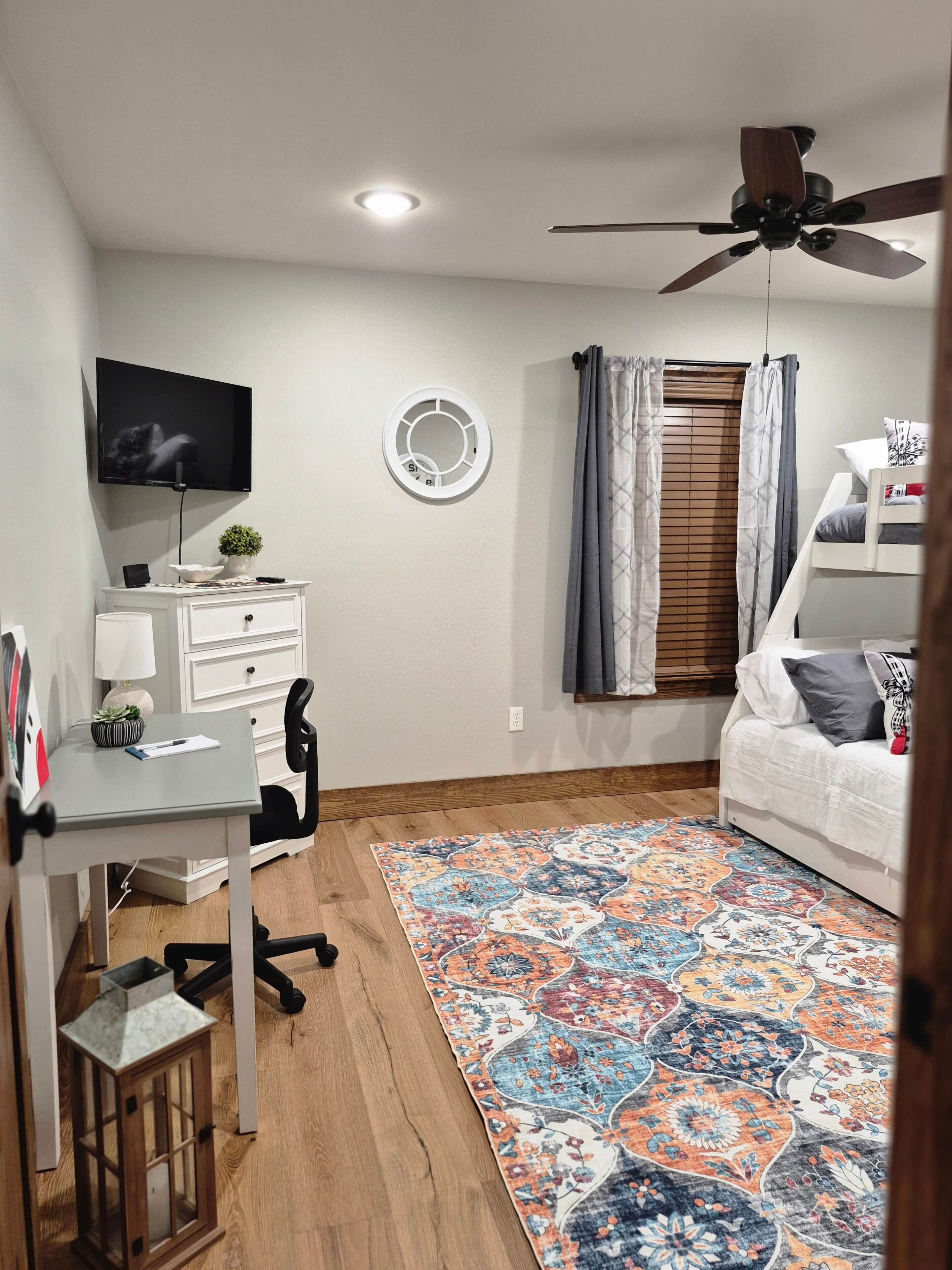 A bedroom with a bed, a desk, a chair, a television, a ceiling fan, a window with curtains, and a patterned rug.