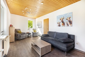 Ein modernes Wohnzimmer mit Holzbalkendecke, einem grauen Sofa, einem Holztisch und einem Bild von Kühen an der Wand.