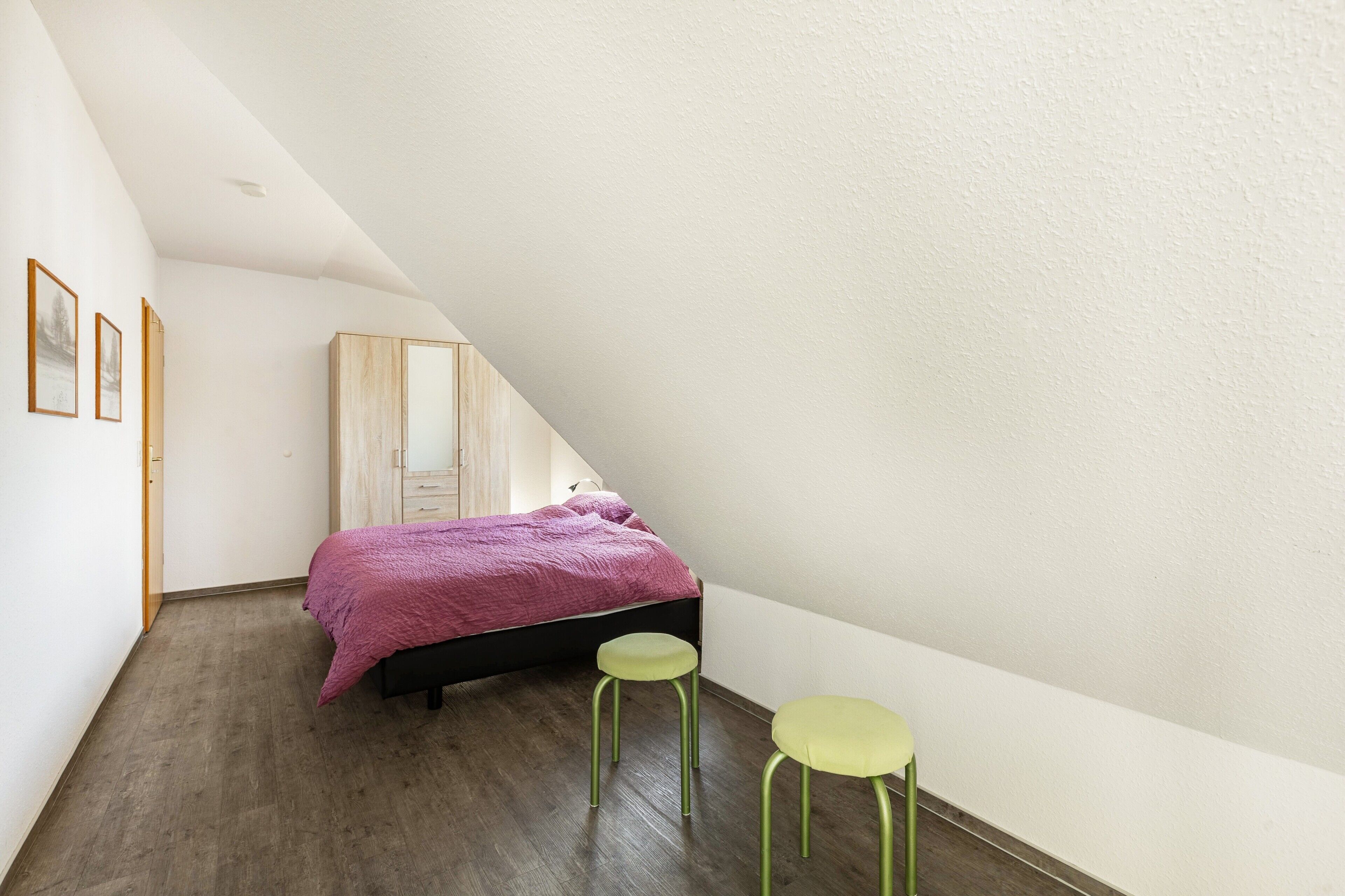 A bedroom with a bed, two stools, a wardrobe, and framed pictures on the wall.