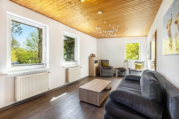 Ein modernes Wohnzimmer mit Holzboden, großen Fenstern, einem grauen Sofa und einem Couchtisch aus Holz.