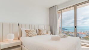 A modern bedroom with a large bed, white bedding, and a view of a cityscape through floor-to-ceiling windows.