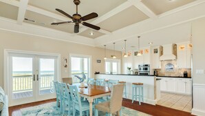 A spacious dining area with a wooden table and blue chairs, a ceiling fan, and a view of the ocean through large sliding doors. - Bright Beachfront Retreat, Elevator, Dog Friendly, Quick Beach Access (Galveston)