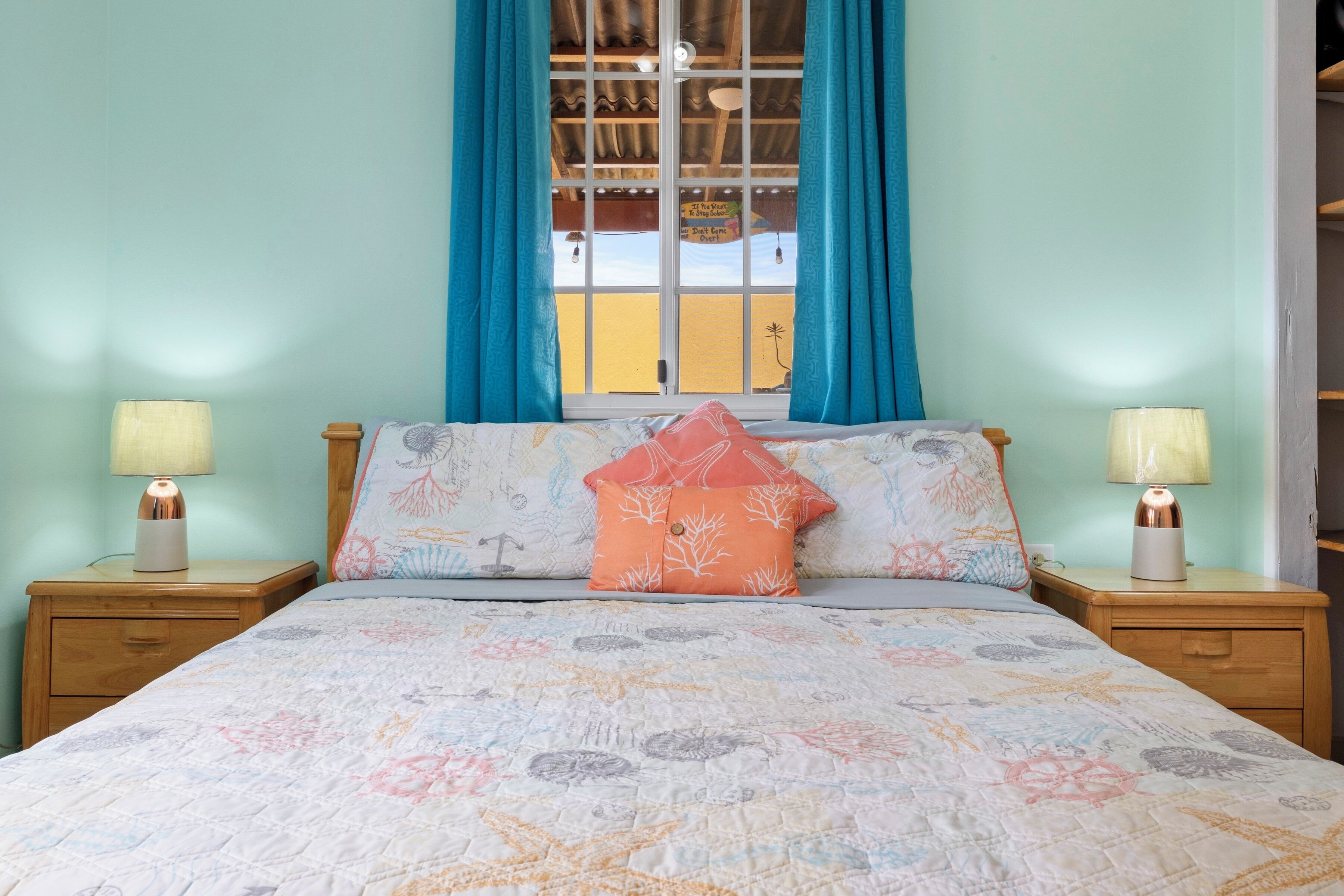 A bedroom with a bed, two bedside tables with lamps, a window with curtains, and a view of the outside.