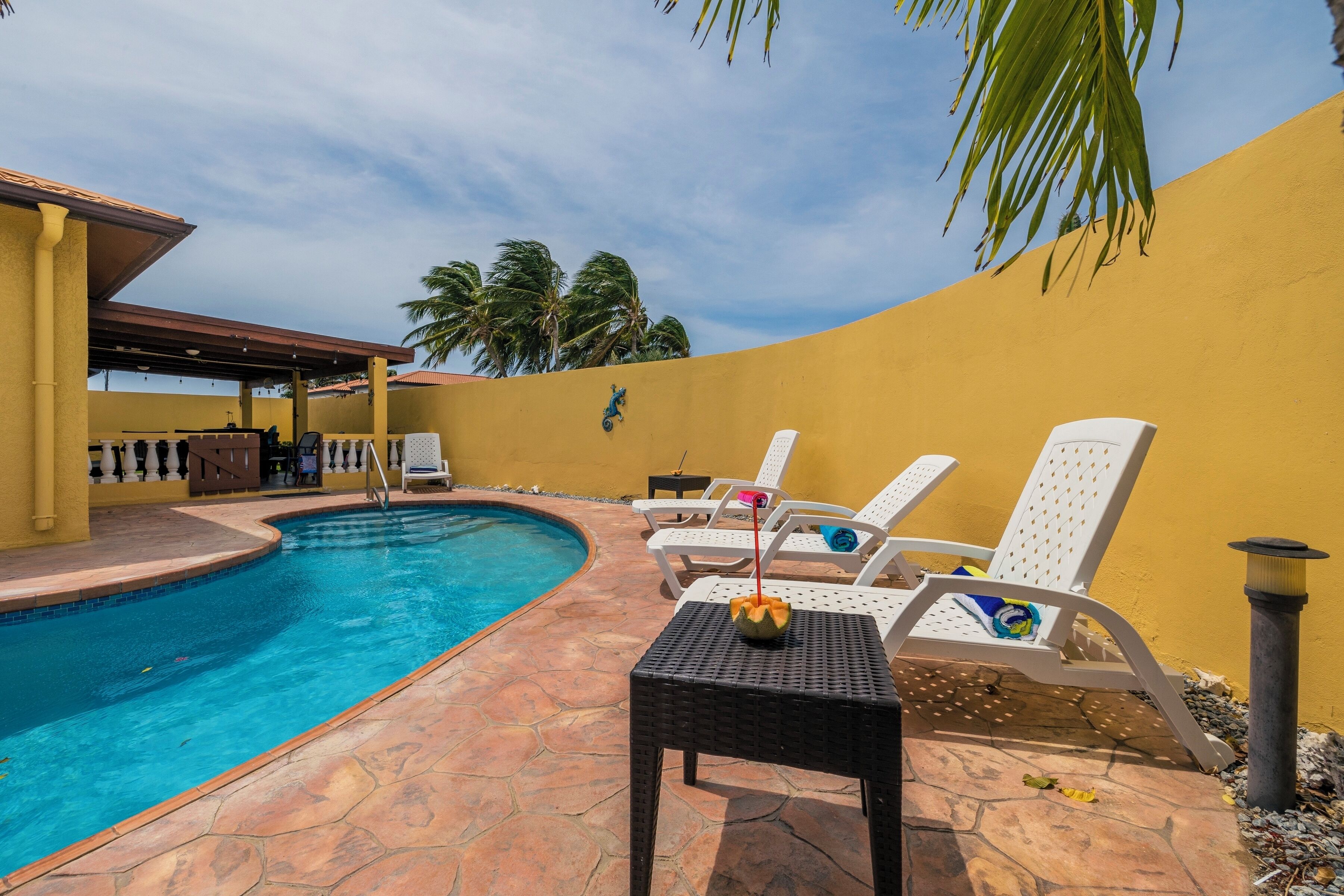 A pool area with lounge chairs and a table, surrounded by a yellow wall and palm trees.