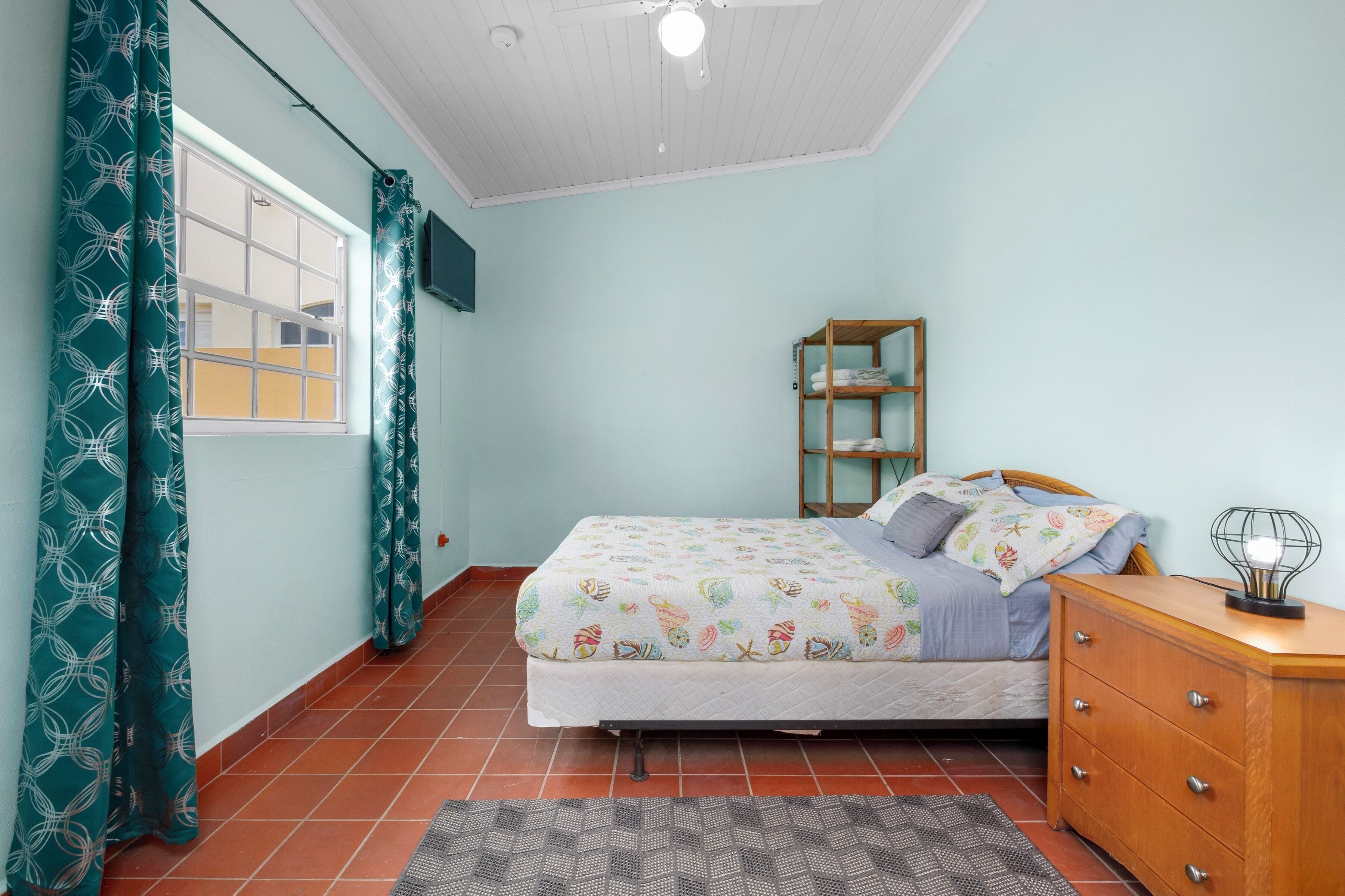 A bedroom with a bed, a wooden dresser, a shelf, and a window with curtains.