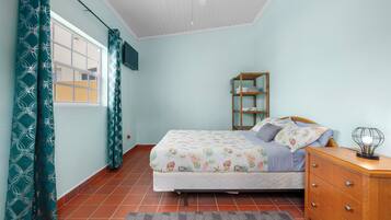 A bedroom with a bed, a wooden dresser, a shelf, and a window with curtains.